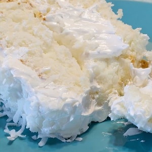 Puede incluir: Primer plano de un pastel blanco con copos de coco en un plato azul. El pastel está cubierto con glaseado blanco y tiene una rebanada cortada.