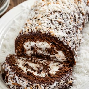 May include: A chocolate cake roll covered in chocolate frosting and shredded coconut. The cake roll is sliced open to reveal a white filling.