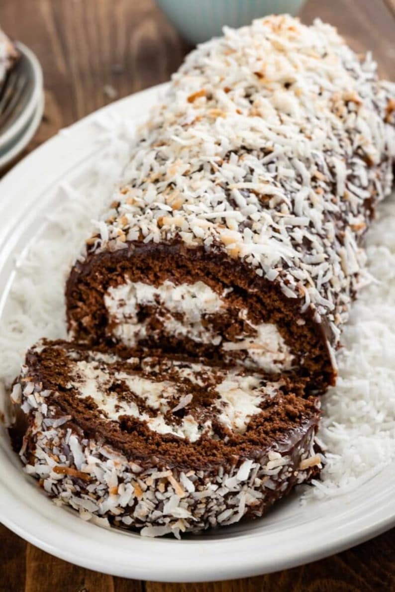 May include: A chocolate cake roll covered in chocolate frosting and shredded coconut. The cake roll is sliced open to reveal a white filling.