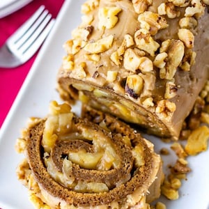 May include: A close-up of a sliced apple cinnamon roll topped with a caramel glaze and chopped walnuts. The roll is on a white plate.