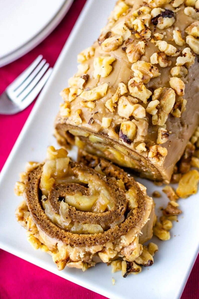 May include: A close-up of a sliced apple cinnamon roll topped with a caramel glaze and chopped walnuts. The roll is on a white plate.