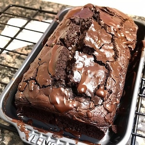May include: A loaf of chocolate cake with a rich chocolate glaze, sitting in a dark gray loaf pan. The cake is cracked and has a slightly uneven surface.