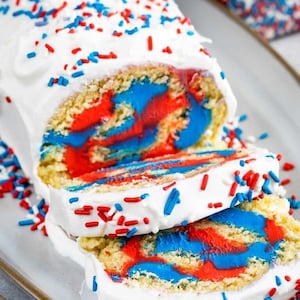 May include: A red, white, and blue swirl cake roll with white frosting and red and blue sprinkles. The cake roll is sliced and ready to be served.