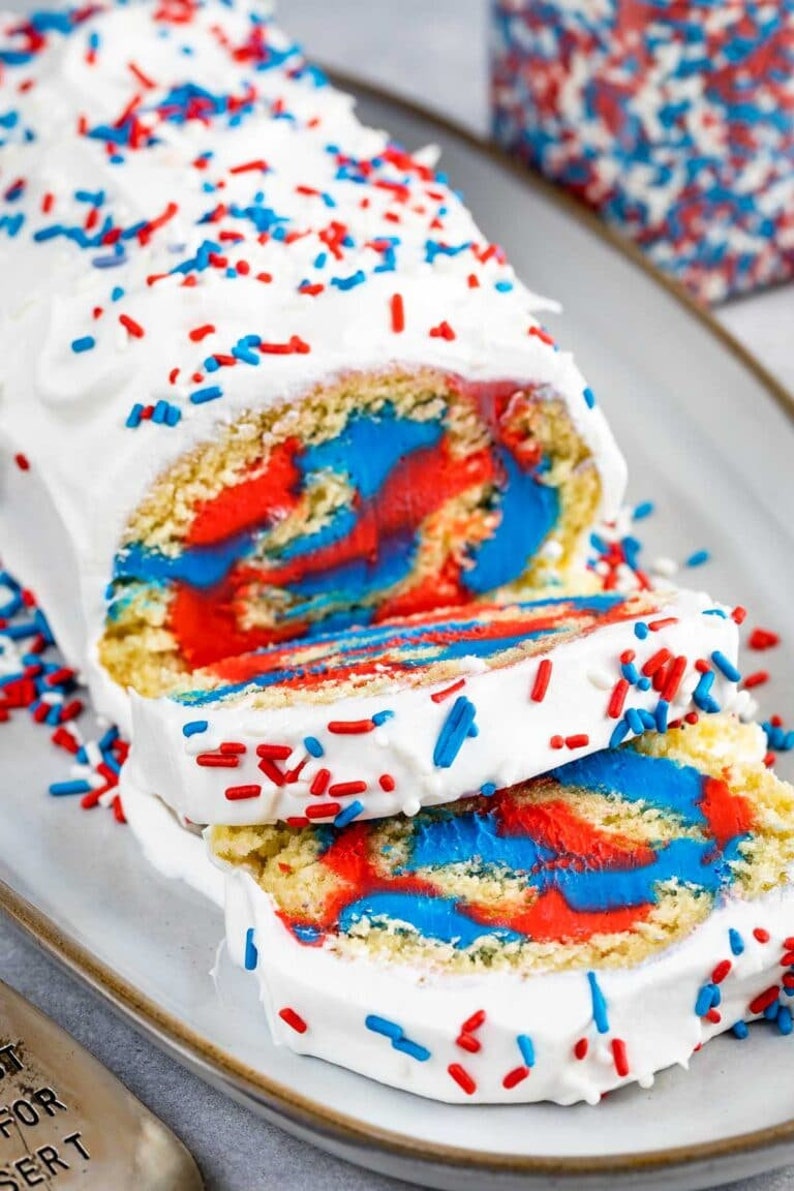May include: A red, white, and blue swirl cake roll with white frosting and red and blue sprinkles. The cake roll is sliced and ready to be served.