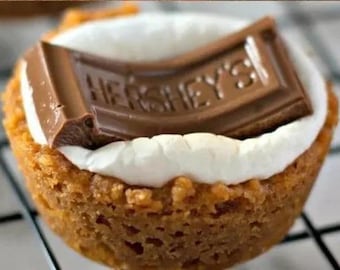 Può includere: Primo piano di un s'more, con una base di biscotto Graham, uno strato di marshmallow bianco e un pezzo di cioccolato Hershey's. La barretta di cioccolato è spezzata, con il logo Hershey's visibile.