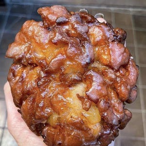 May include: A golden-brown apple fritter, deep-fried and glistening. Its irregular, craggy surface shows a rich, caramelized texture. The fritter's color ranges from light to dark brown, indicating varying degrees of browning.  The overall appearance suggests a freshly made, warm, and possibly sweet treat. The fritter's size is substantial, filling the frame.