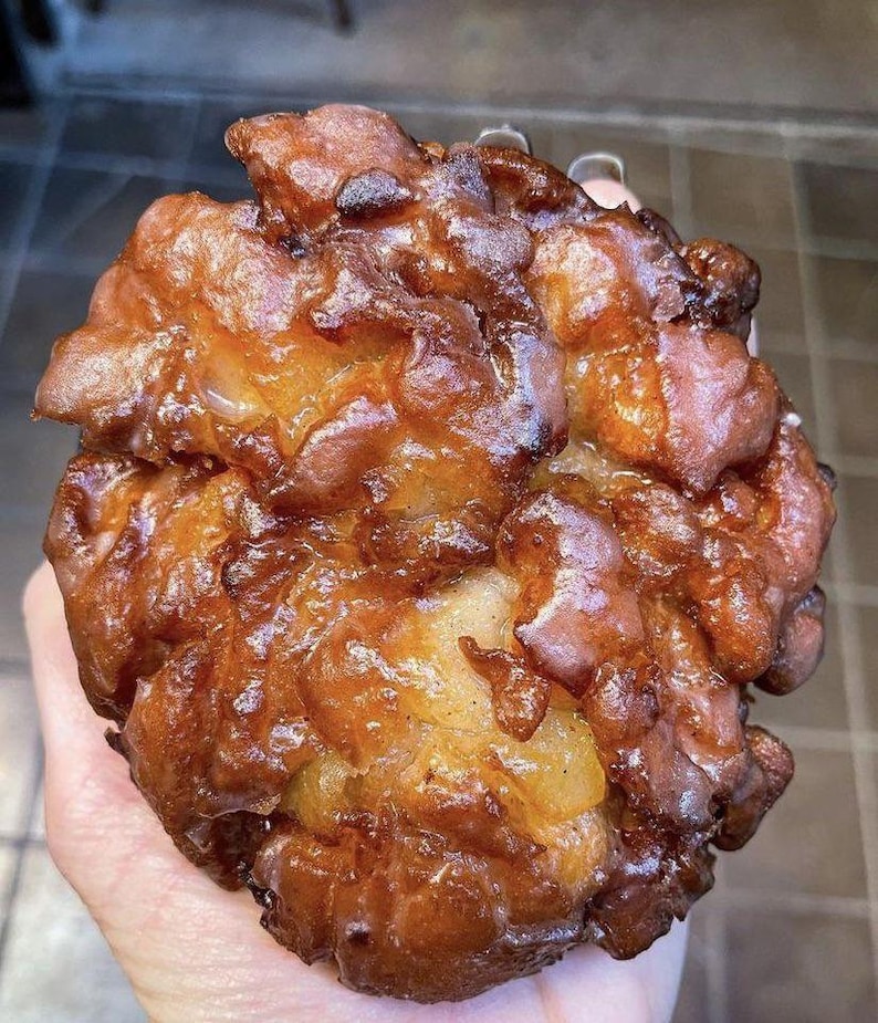 May include: A golden-brown apple fritter, deep-fried and glistening. Its irregular, craggy surface shows a rich, caramelized texture. The fritter's color ranges from light to dark brown, indicating varying degrees of browning.  The overall appearance suggests a freshly made, warm, and possibly sweet treat. The fritter's size is substantial, filling the frame.