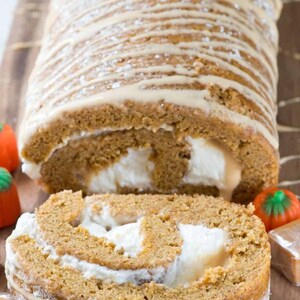May include: A pumpkin spice roll cake with a white cream filling and a caramel glaze. The cake is dusted with powdered sugar and is sliced in half, revealing the filling. The cake is on a wooden cutting board with candy pumpkins and caramel candies.