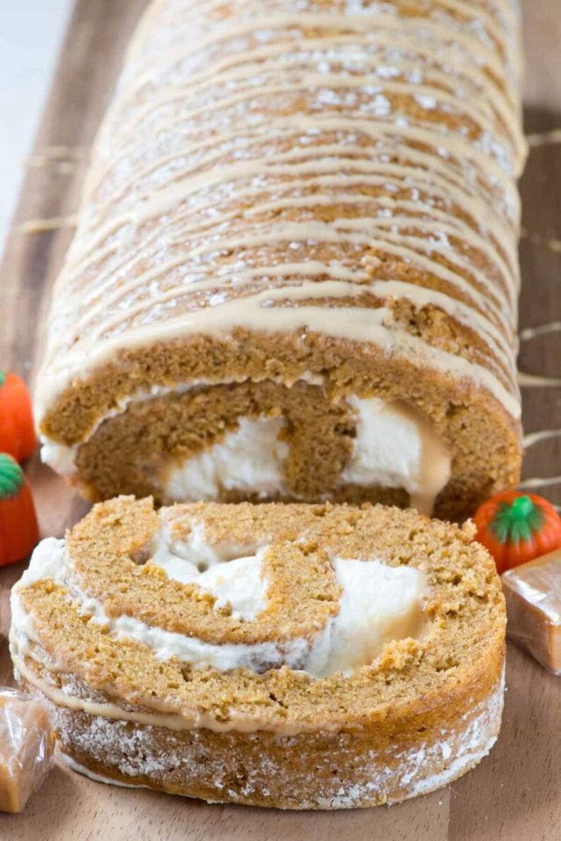 May include: A pumpkin spice roll cake with a white cream filling and a caramel glaze. The cake is dusted with powdered sugar and is sliced in half, revealing the filling. The cake is on a wooden cutting board with candy pumpkins and caramel candies.