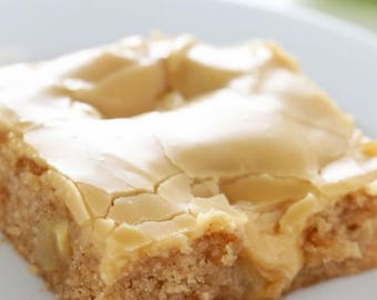 Op de afbeelding: Een close-up van een vierkant stuk cake met lichtbruine frosting. De frosting heeft een gebarsten uiterlijk en een licht onregelmatig oppervlak. De cake is lichtbruin van kleur en heeft een licht kruimelige textuur.