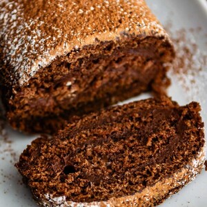 May include: A chocolate cake roll dusted with powdered sugar, sliced into a single piece. The cake is a rich brown color and has a soft, fluffy texture.