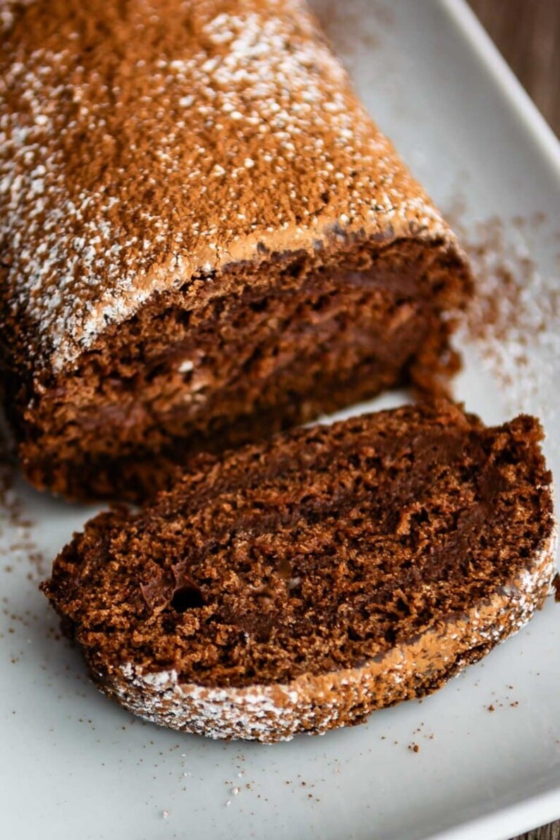 May include: A chocolate cake roll dusted with powdered sugar, sliced into a single piece. The cake is a rich brown color and has a soft, fluffy texture.