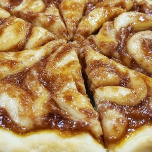 May include: A close-up of a cinnamon roll pizza cut into slices. The pizza is topped with a brown cinnamon swirl and a white icing. The crust is golden brown.