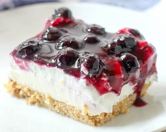 May include: A slice of blueberry cheesecake dessert with a graham cracker crust, topped with a blueberry sauce and fresh blueberries.