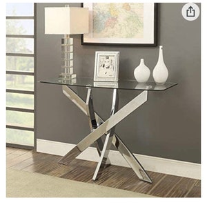 Puede incluir: Una mesa de sofá de metal cromado plateado en forma de X con una tapa de vidrio transparente. La mesa tiene un diseño contemporáneo de transición e incluye herrajes. La mesa se muestra con una lámpara, un marco de imagen y dos jarrones blancos.
