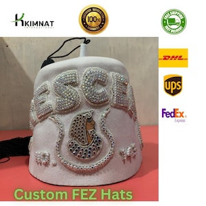 Puede incluir: Un sombrero fez blanco con la palabra "ESCEA" en pedrería brillante. El sombrero presenta un diseño bordado de un faraón y el texto "Custom FEZ Hats" en verde.