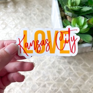 Può includere: Un adesivo bianco con le parole "Love Kansas City" in lettere gialle e rosse. L'adesivo è tenuto da una mano.
