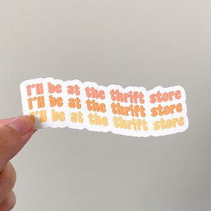 May include: A white sticker with the text "I'll be at the thrift store" in orange and yellow bubble letters. The sticker is held by a hand against a neutral background.