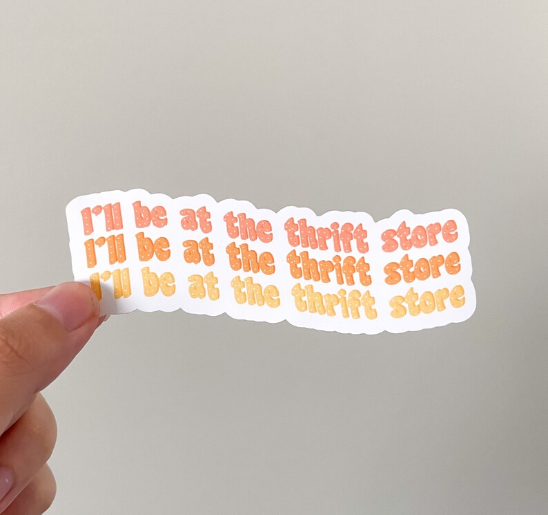 May include: A white sticker with the text "I'll be at the thrift store" in orange and yellow bubble letters. The sticker is held by a hand against a neutral background.