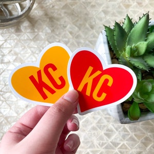 May include: Two heart-shaped stickers with the letters "KC" in white on a red and yellow background.