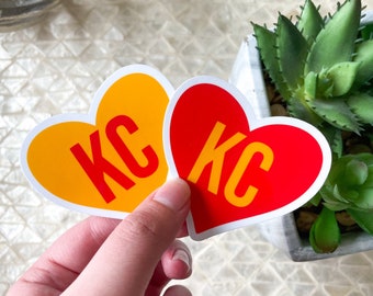 Può includere: Due adesivi a forma di cuore con le lettere "KC" in bianco su sfondo rosso e giallo.