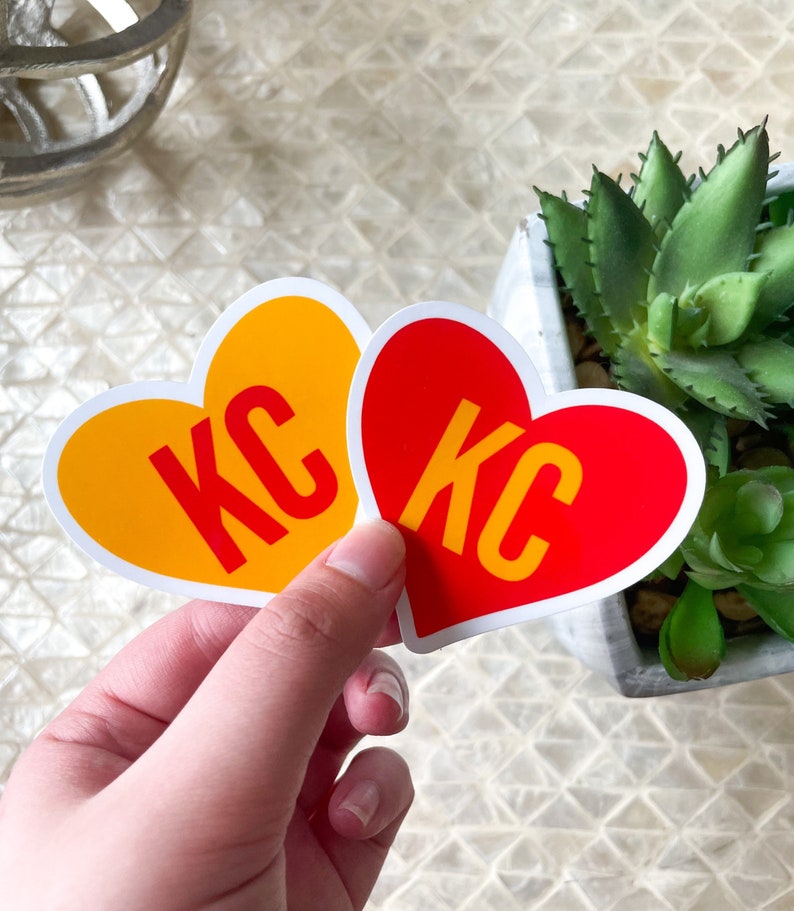 May include: Two heart-shaped stickers with the letters "KC" in white on a red and yellow background.