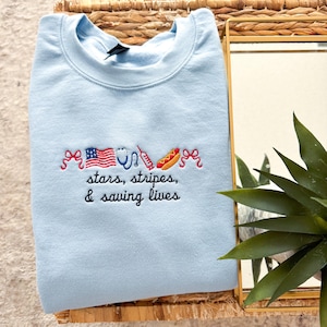 May include: A light blue crewneck sweatshirt featuring embroidered designs of red bows, an American flag, a stethoscope, a syringe, and a hot dog. Below the icons, the text reads, "stars, stripes, & saving lives" in black cursive lettering.