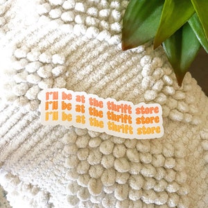 May include: Three stickers with the text "I'll be at the thrift store" in orange and yellow, on a white textured surface. The stickers are arranged in a row. A green plant is visible in the background.