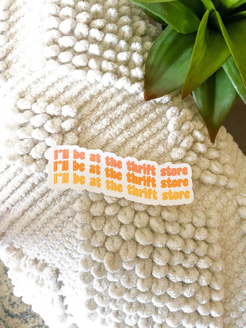 May include: Three stickers with the text "I'll be at the thrift store" in orange and yellow, on a white textured surface. The stickers are arranged in a row. A green plant is visible in the background.