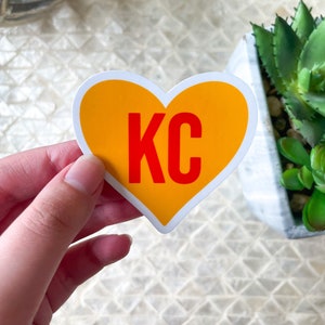 May include: A yellow heart-shaped sticker with the letters "KC" in red inside. The sticker is being held by a hand.
