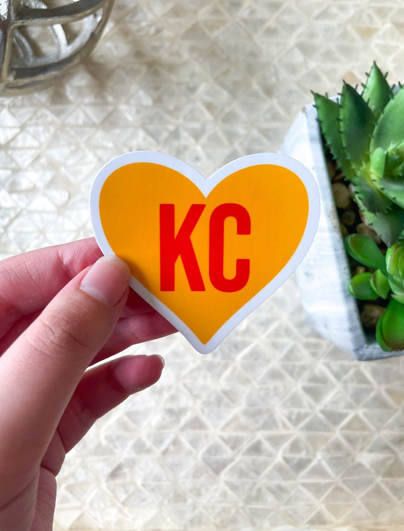 May include: A yellow heart-shaped sticker with the letters "KC" in red inside. The sticker is being held by a hand.