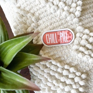 May include: A pink and white pill-shaped sticker with the text "CHILL PILL" on a white fuzzy surface.
