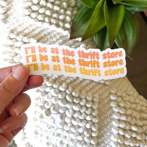 May include: A sticker with the text "I'll be at the thrift store" in orange and yellow. The sticker is held by a hand against a white textured background, with a green plant in the background.