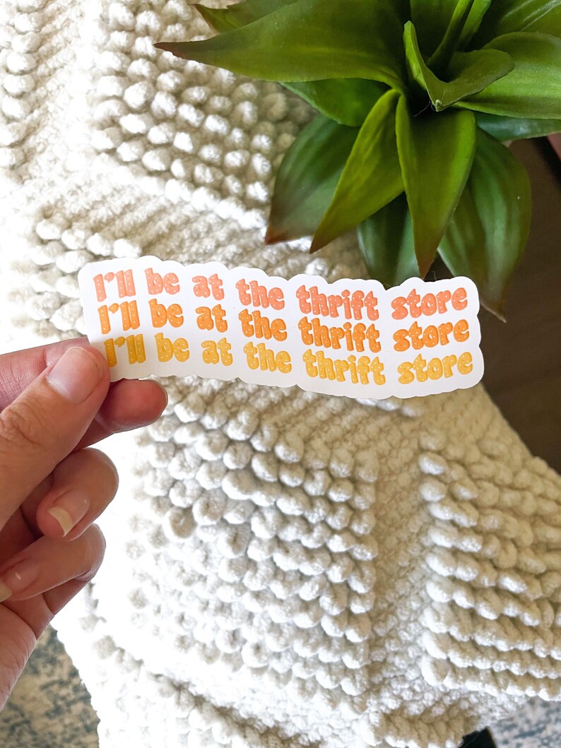 May include: A sticker with the text "I'll be at the thrift store" in orange and yellow. The sticker is held by a hand against a white textured background, with a green plant in the background.