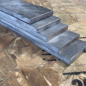 May include: Five pieces of flat, rectangular metal bars stacked on top of each other. The bars are a dark gray color and have a smooth, polished finish. The bars are arranged in a graduated size, with the largest bar at the bottom and the smallest bar at the top.
