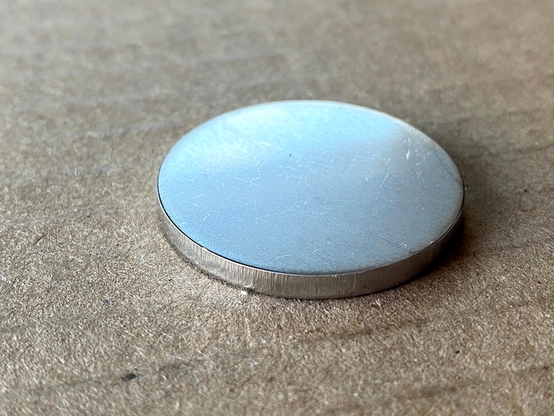 May include: A silver-coloured metal disc with a smooth, slightly textured surface. The disc has a slightly raised edge.