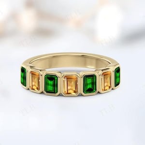May include: A gold ring featuring alternating emerald green and amber-colored rectangular gemstones. The ring has a smooth, polished gold band and a series of bezel-set stones. The gemstones are evenly spaced around the ring.