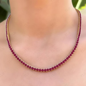 Puede incluir: Un collar de cadena de oro con una fila de pequeñas piedras preciosas redondas rojas. El collar está en el cuello de una modelo.
