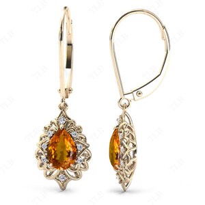 Op de afbeelding: Een paar gouden oorhangers met een peervormige oranje edelsteen omringd door kleinere witte edelstenen. De oorbellen hebben een sluiting met een hendel.