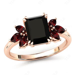 May include: A rose gold ring featuring a large black square-shaped gemstone and smaller red gemstones set into the band.