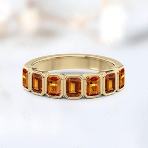 Puede incluir: Un anillo de oro con una serie de piedras preciosas rectangulares de color naranja-amarillo. Las piedras están engastadas en un estilo biselado, creando una banda continua de color. El anillo tiene un acabado pulido y liso.