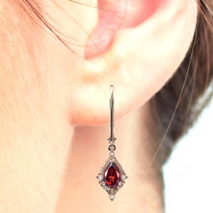 May include: A pair of silver dangle earrings with a pear-shaped red gemstone surrounded by smaller white gemstones.