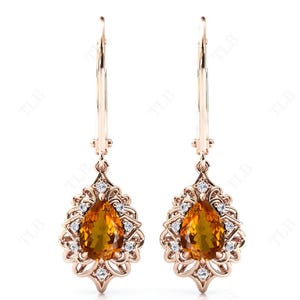 Op de afbeelding: Een paar gouden oorhangers met peervormige oranje edelstenen, omringd door kleinere heldere edelstenen.