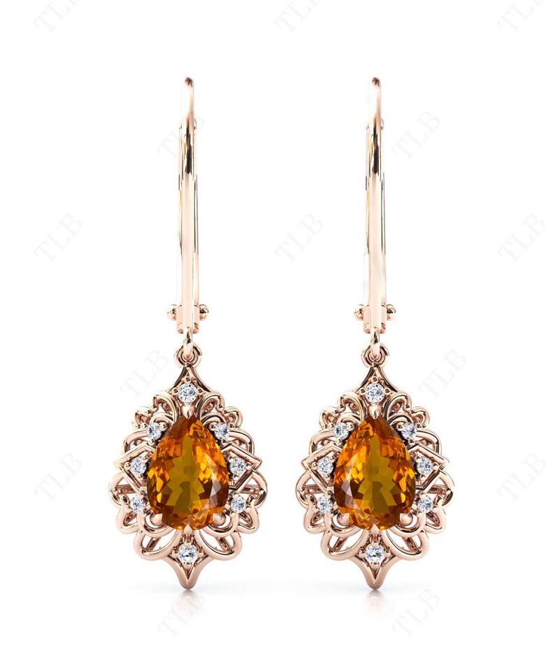 Op de afbeelding: Een paar gouden oorhangers met peervormige oranje edelstenen, omringd door kleinere heldere edelstenen.