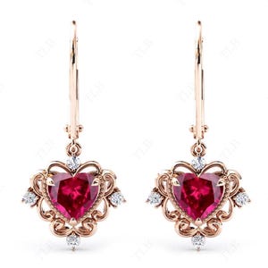 Puede incluir: Un par de pendientes de corazón de oro rosa con una gran piedra preciosa de rubí rojo en el centro de cada corazón. Los corazones están rodeados de piedras preciosas más pequeñas y transparentes.
