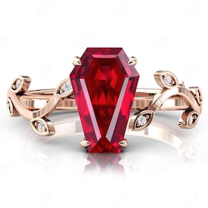 Puede incluir: Un anillo de oro rosa con una gran piedra preciosa facetada de color rojo oscuro engastada en forma de ataúd. La banda es un delicado diseño de vid con pequeños diamantes engastados en las hojas.