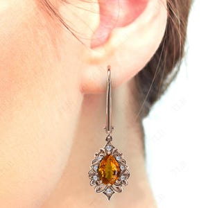 Op de afbeelding: Een paar gouden oorhangers met een peervormige oranje edelsteen omringd door kleinere heldere edelstenen.