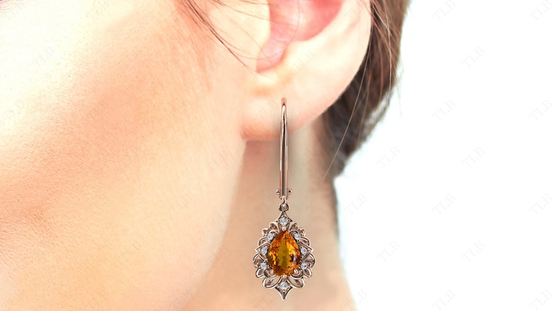 Op de afbeelding: Een paar gouden oorhangers met een peervormige oranje edelsteen omringd door kleinere heldere edelstenen.