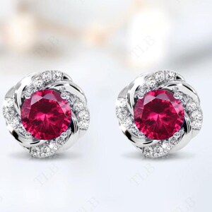 May include: A pair of silver earrings with a red gemstone surrounded by a halo of smaller white gemstones.