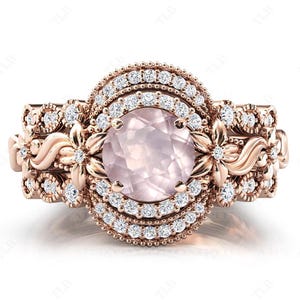 Könnte beinhalten: Ein Verlobungsring aus Roségold mit einem großen ovalen rosafarbenen Edelstein, der von einem Heiligenschein aus kleineren Diamanten umgeben ist. Der Ring hat komplizierte florale Details und eine zarte Band.